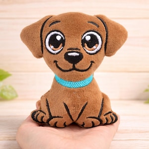 May include: A brown plush puppy with large, expressive eyes and a blue collar. The stuffed animal has a smiling face and black paws. The toy is held in a person's hand against a wooden background.