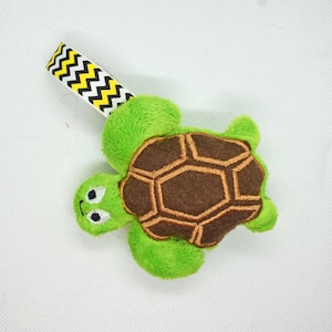 May include: A plush green turtle toy with a brown shell and embroidered details. The turtle has a cheerful expression and is attached to a white ribbon with a black and yellow zigzag pattern. A cute, soft toy.