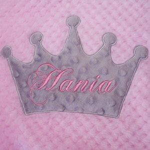 May include: A pink, textured blanket with a gray crown applique. The crown is embroidered with the name "Hania" in pink script. The blanket has a soft, plush appearance.