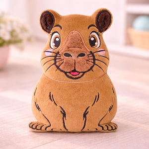May include: A plush, brown capybara stuffed animal with embroidered details. The toy features a smiling face with pink cheeks, black whiskers, and a small, open mouth. The capybara has small, rounded ears and is sitting upright.