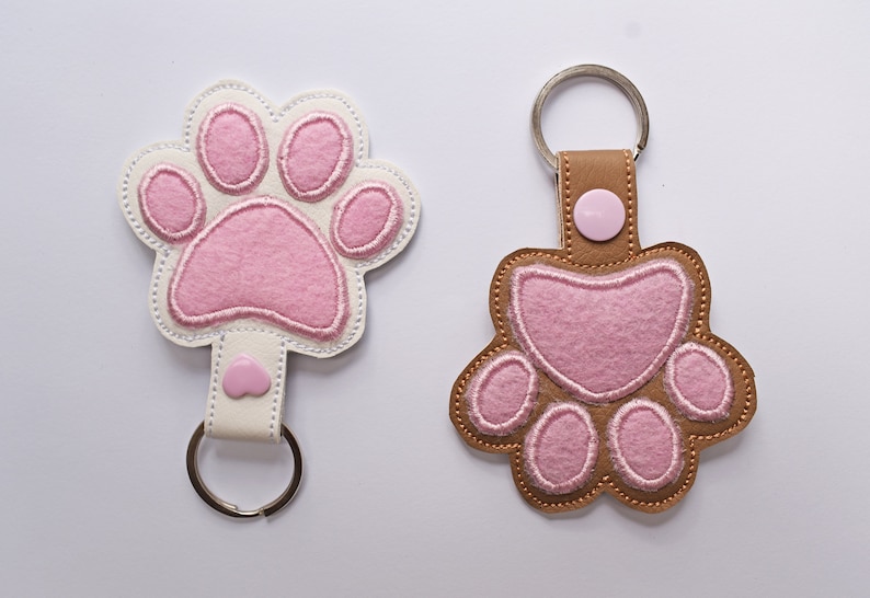 May include: Two keychains in the shape of a paw print. One is white with pink accents and a silver key ring. The other is brown with pink accents and a silver key ring. Both have a button closure.