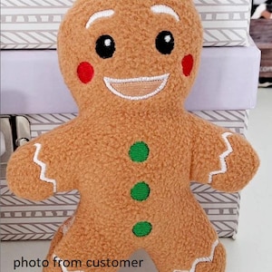 Op de afbeelding: Een zacht gingerbread man speelgoed met een lachend gezicht, zwarte ogen en rode wangen. De gingerbread man heeft groene knoopaccenten en witte glazuurdetails. De tekst "photo from customer" is zichtbaar onderaan de afbeelding.