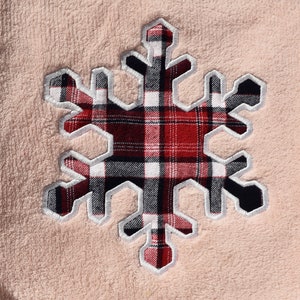 May include: A close-up of a peach-coloured fabric with a large snowflake appliqué. The snowflake has a red, black, and white plaid pattern, outlined with silver-grey stitching. The design is centred on the fabric.