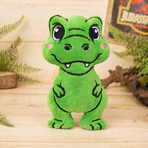 May include: A plush green dinosaur toy with black outlines, large eyes, and pink cheeks. The dinosaur is standing on a wooden surface. The background includes ferns and a Jurassic Park sign.