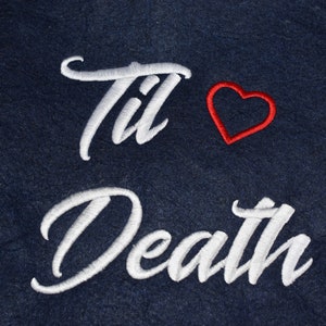 Könnte beinhalten: Dunkelblauer Stoff mit weißer Stickerei "Til Death" und einem roten Herzsymbol. Die geschwungene Stickschrift und das rote Herz heben sich vom dunklen Hintergrund ab.
