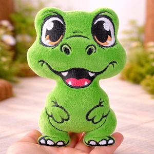 May include: A green plush frog toy with large eyes, a smiling mouth, and small limbs. The frog has a pink tongue and white teeth. The toy is held in a hand, with a blurred background of greenery.