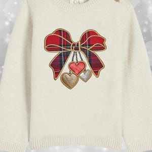 Puede incluir: Suéter color crema con un diseño decorativo de lazo y corazones. El lazo presenta un patrón de cuadros rojos y verdes con ribetes dorados. Tres corazones colgantes en rojo, plateado y dorado completan el diseño. El suéter tiene mangas largas y un dobladillo acanalado.