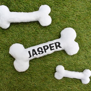 Puede incluir: Juguetes de hueso para perros de felpa blanca sobre hierba verde. Un hueso tiene el nombre "JASPER" bordado en negro. Los otros dos huesos son lisos. Estos juguetes blandos son para mascotas.