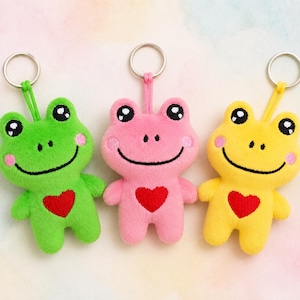 Op de afbeelding: Drie pluche kikker sleutelhangers in groen, roze en geel. Elke kikker heeft een lachend gezicht, roze wangen en een rood hart op de borst. De sleutelhangers hebben zilveren ringen en staan tegen een zachte, aquarel achtergrond.