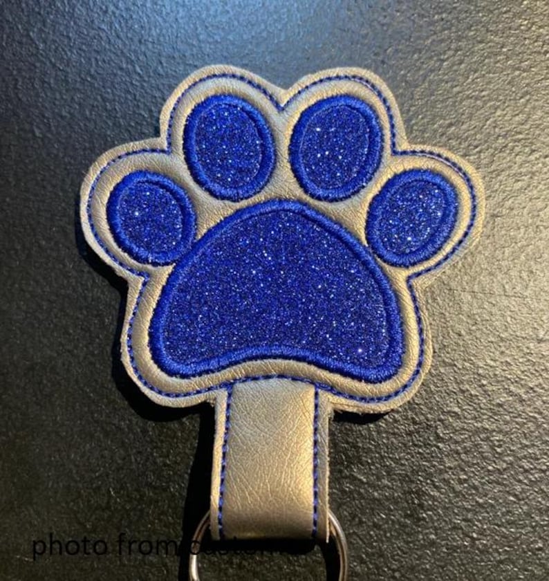 May include: A blue glitter paw print keychain with a gold border and a gold leather strap. The paw print is stitched onto the gold leather strap. The keychain has a silver ring.