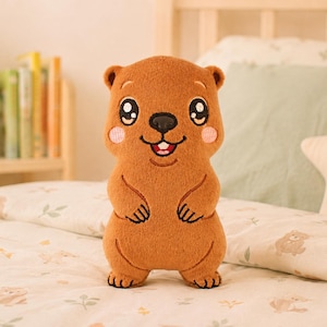 May include: A brown plush toy shaped like a quokka with embroidered facial features, including large black eyes with white highlights, a black nose, a smiling mouth with visible teeth, and pink circular cheeks, standing upright on a soft surface.