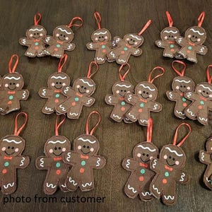May include: A collection of brown felt gingerbread man ornaments with white icing details, red bow ties, and red ribbon hangers. Each ornament has a smiling face and decorative buttons. The phrase "photo from customer" is visible.