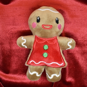 May include: A plush gingerbread person doll with a red dress and white icing details. The doll has pink cheeks, black eyes, and a smiling mouth. The background is a deep red fabric.