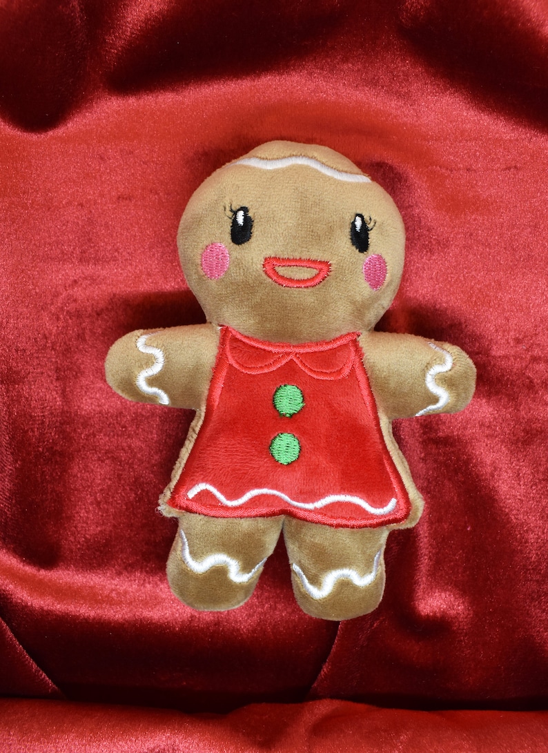 May include: A plush gingerbread person doll with a red dress and white icing details. The doll has pink cheeks, black eyes, and a smiling mouth. The background is a deep red fabric.