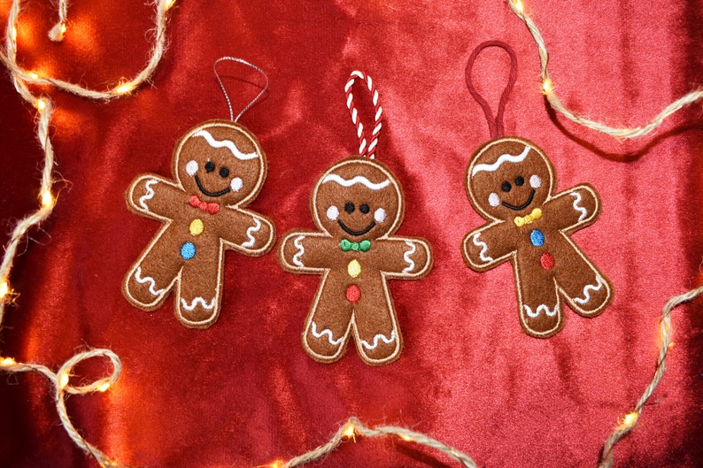 May include: Three gingerbread man ornaments with white icing details, colorful buttons, and smiling faces. Each ornament has a different colored bow tie and a loop for hanging. Set against a red background with string lights.