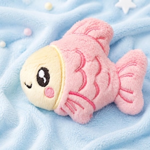 May include: A plush fish toy featuring a soft cream-colored face with embroidered black eyes and pink cheeks, attached to a textured pink body with stitched scale details and fins. The toy has a rounded shape and a fuzzy, soft-touch fabric finish.