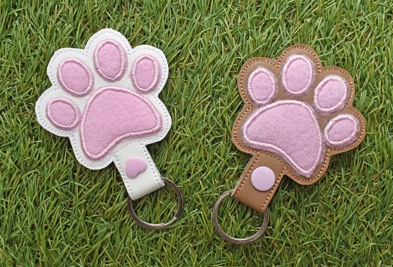 May include: Two paw-shaped keychains. One is white with a pink paw print, the other is brown with a pink paw print. Both have a metal keyring and a small heart-shaped accent. The keychains are on a green grass background.