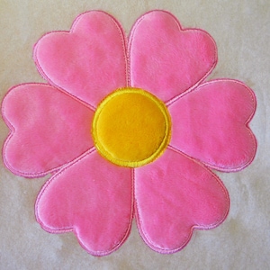 Könnte beinhalten: Eine plüschige rosa Blume mit herzförmigen Blütenblättern und einem gelben Zentrum. Die Blume ist mit einem gelben Umriss bestickt und liegt auf einem cremefarbenen Hintergrund. Dieses dekorative Blumendesign eignet sich für verschiedene Bastelprojekte.