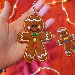 May include: Two gingerbread man ornaments. The larger ornament is held in a hand and has white icing details, a green bow tie, and colorful buttons. The smaller ornament is in the background. Both have red and white string for hanging.