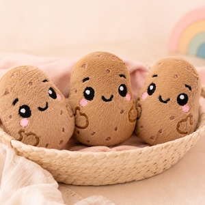 Op de afbeelding: Drie pluche aardappelspeeltjes met geborduurde gezichtjes, voorzien van grote zwarte ogen, kleine glimlachjes en roze wangen. Elke bruine, getextureerde aardappel heeft kleine geborduurde armpjes en donkerbruine vlekjes in een gevlochten mand.