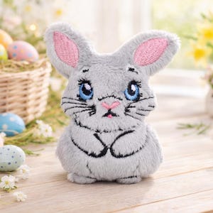 May include: A plush gray bunny with pink inner ears, blue eyes, and black whiskers. The bunny is a decorative item, possibly for Easter. The background includes a basket of decorated eggs and flowers.