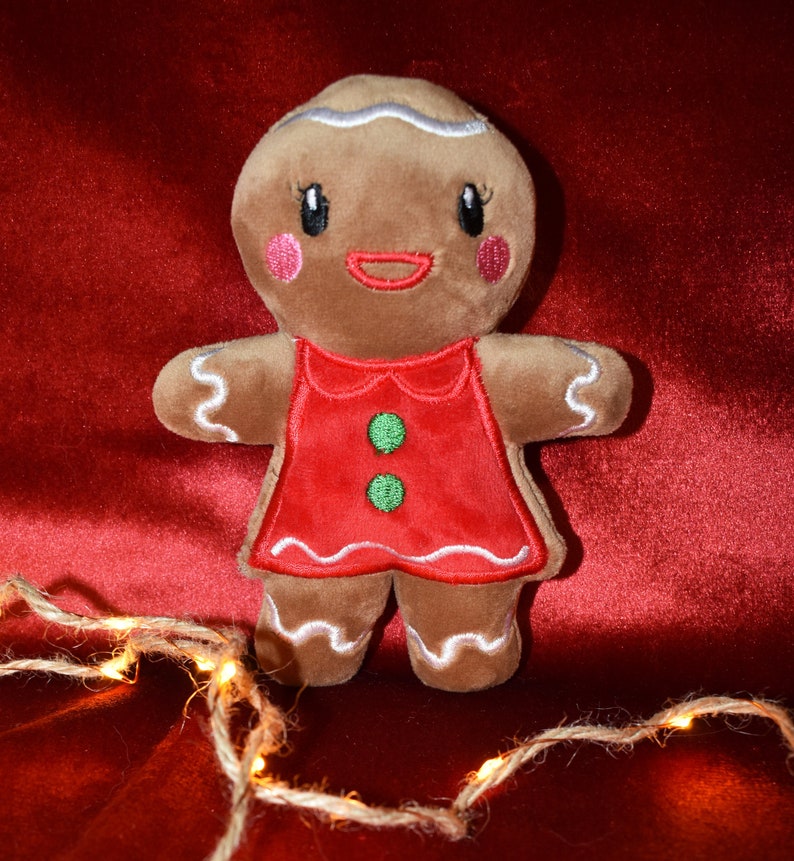 May include: A plush gingerbread person toy with a red dress and green buttons. The gingerbread person has white icing details, pink cheeks, and a smiling red mouth. The toy is set against a red velvet background, with a string of lights in the foreground.