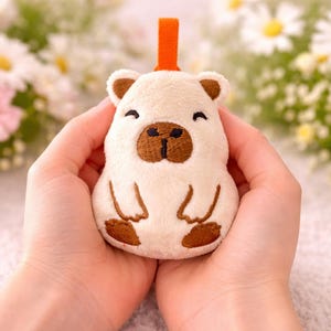 May include: A plush, cream-colored capybara toy with brown accents, including the ears, nose, and feet. The toy has a small orange loop at the top. The capybara is held in two hands, with a floral background.