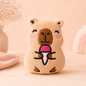 Peut inclure: Une peluche capybara beige avec des détails brodés, incluant un museau marron foncé, des yeux fermés, des joues roses et de petits pieds. Le capybara tient un petit cornet de glace avec une boule rose vif et un cornet blanc dans ses pattes.