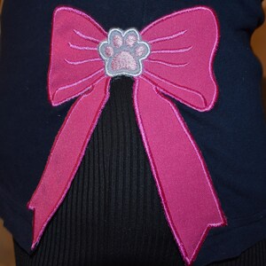 May include: Navy blue pet shirt with a large pink bow applique. The bow features a paw print in the center. The shirt is made of a soft material and is designed for pets.