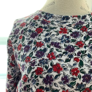 May include: A long-sleeved top with a floral design. The garment has a white base covered in a pattern of red, purple, and blue flowers with green foliage. The fabric has a ribbed texture.
