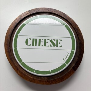 May include: A round wooden cheese board with a white ceramic insert. The insert features the word "CHEESE" in green block letters. A green border surrounds the white insert. The board is a food serving accessory.