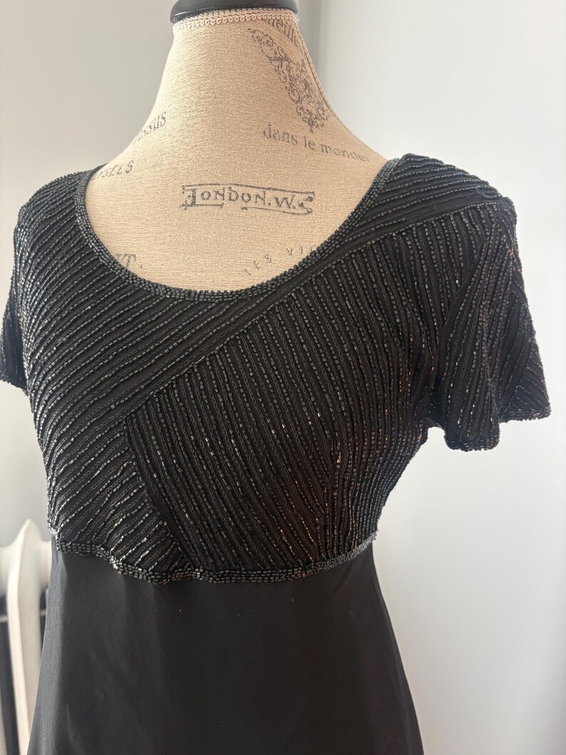 Peut inclure: Robe noire avec un corsage perl&eacute; et des manches courtes. Le corsage pr&eacute;sente des motifs perl&eacute;s diagonaux et horizontaux. La robe a une encolure d&eacute;gag&eacute;e et une jupe noire unie. La robe est pr&eacute;sent&eacute;e sur un mannequin.