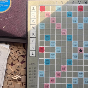 Pode incluir: Um conjunto de jogo de tabuleiro Scrabble vintage. O tabuleiro é cinza com quadrados de pontuação rosa e azul. O conjunto inclui peças de letras de madeira e uma caixa marrom com o logotipo do jogo. O tabuleiro mede cerca de 38 cm quadrados.