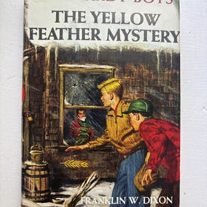 May include: A vintage hardcover book titled The Hardy Boys The Yellow Feather Mystery by Franklin W. Dixon. The cover illustration depicts two figures in a snowy setting looking toward a window where a third figure is visible inside a dark building.