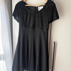 Peut inclure: Une robe noire &agrave; manches courtes est suspendue &agrave; un cintre en bois. Le corsage est orn&eacute; de rang&eacute;es de perles noires et la jupe est en tissu noir uni et fluide. La robe a une encolure d&eacute;gag&eacute;e et un ourlet festonn&eacute;.
