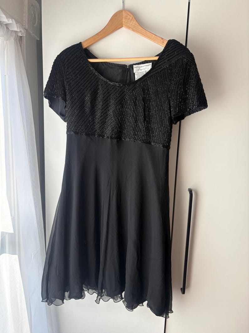 Peut inclure: Une robe noire &agrave; manches courtes est suspendue &agrave; un cintre en bois. Le corsage est orn&eacute; de rang&eacute;es de perles noires et la jupe est en tissu noir uni et fluide. La robe a une encolure d&eacute;gag&eacute;e et un ourlet festonn&eacute;.