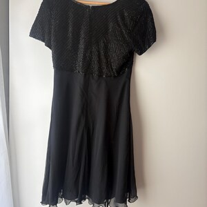 Peut inclure: Robe noire avec un corsage orn&eacute; de perles et des manches courtes. La jupe est en tissu noir uni et fluide, avec un ourlet festonn&eacute;. La robe est suspendue &agrave; un cintre en bois.