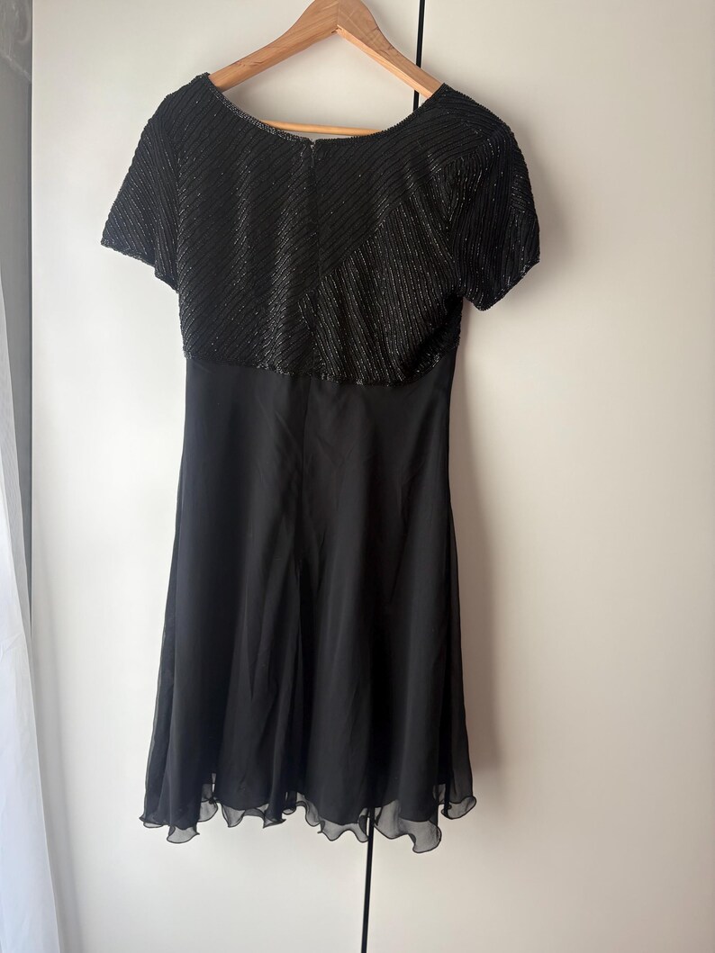Peut inclure: Robe noire avec un corsage orn&eacute; de perles et des manches courtes. La jupe est en tissu noir uni et fluide, avec un ourlet festonn&eacute;. La robe est suspendue &agrave; un cintre en bois.