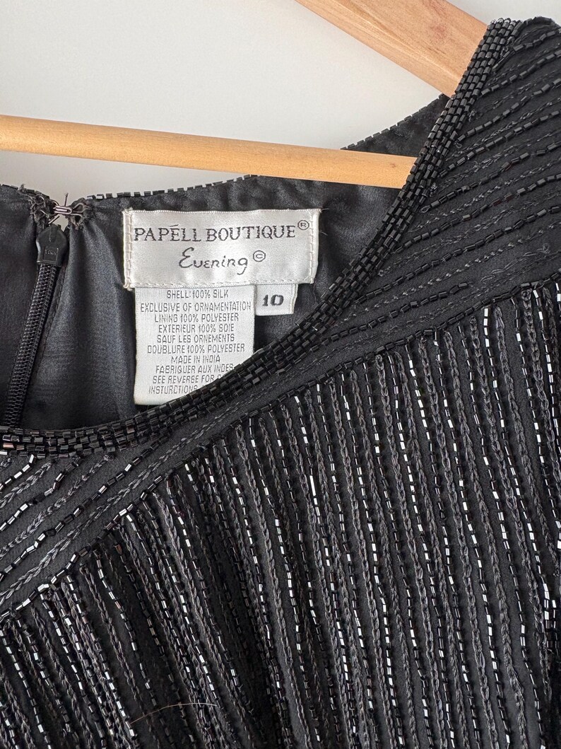 Peut inclure: Gros plan d'une robe de soir&eacute;e noire avec des franges perl&eacute;es et un d&eacute;collet&eacute; perl&eacute;. La robe porte une &eacute;tiquette &laquo; PAPELL BOUTIQUE Evening &raquo; et la taille 10. La robe est en soie et polyester.