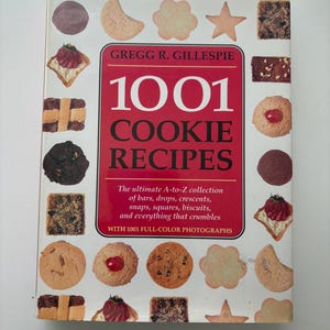以下が含まれることがあります： グレッグ・R・ギレスピー著のハードカバー料理本『1001 Cookie Recipes』。白い表紙の中央には白い文字が書かれた赤い長方形の枠があり、その周囲には星型、円形、四角形など、さまざまなカラフルなクッキーの写真が並んでいます。
