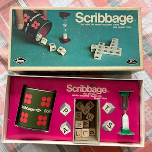 May include: Scribbage word building game set featuring a green shaker cup with red circular patterns, a green and white hourglass timer, and multiple lettered wooden cubes. The game box displays the title Scribbage and the Lowe brand logo.
