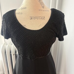 Peut inclure: Robe noire avec un corsage perl&eacute; et des manches courtes. Le corsage pr&eacute;sente un motif g&eacute;om&eacute;trique de perles noires. La robe est pr&eacute;sent&eacute;e sur un mannequin.