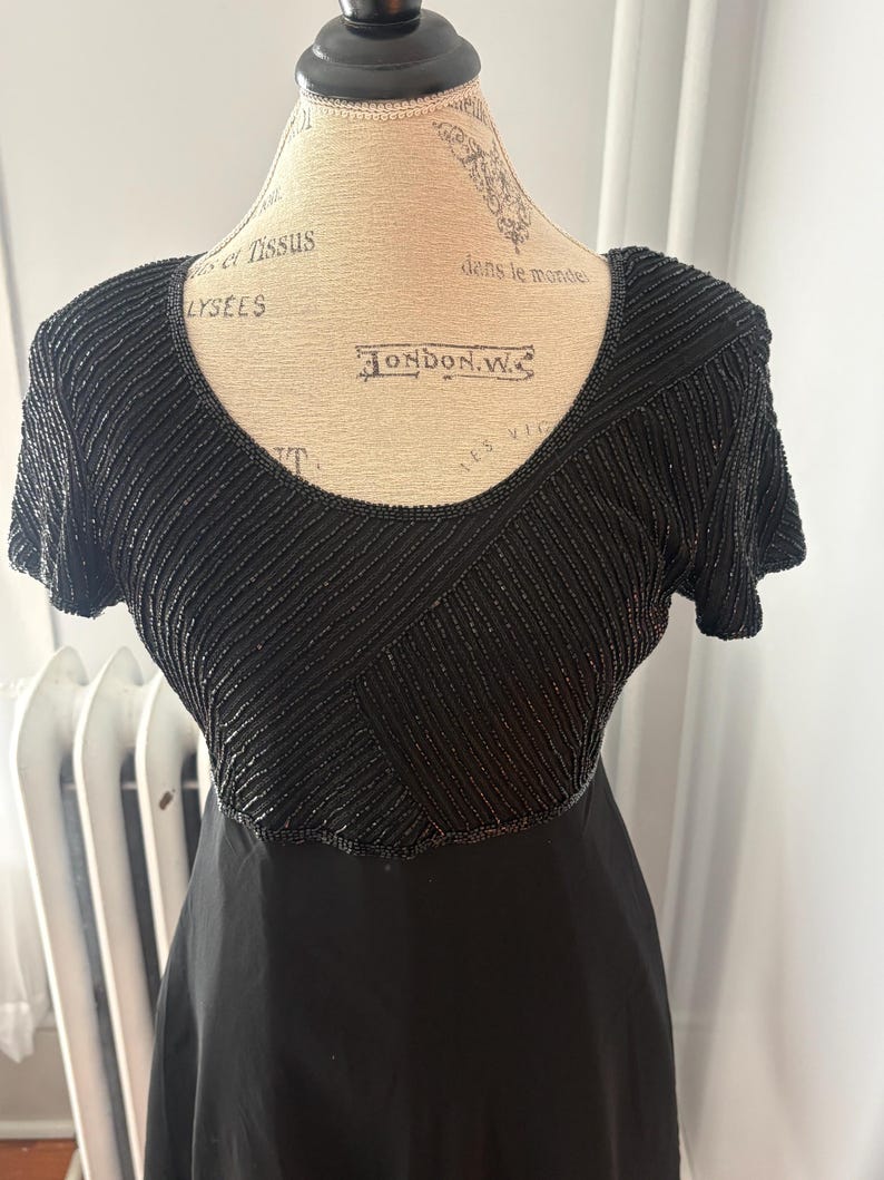 Peut inclure: Robe noire avec un corsage perl&eacute; et des manches courtes. Le corsage pr&eacute;sente un motif g&eacute;om&eacute;trique de perles noires. La robe est pr&eacute;sent&eacute;e sur un mannequin.