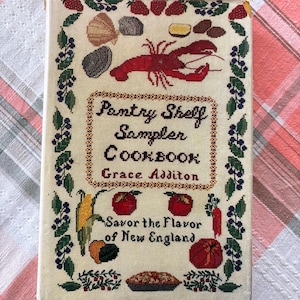 May include: A hardback cookbook titled Pantry Shelf Sampler Cookbook by Grace Additon, featuring a cream-coloured cover with cross-stitch embroidery of a red lobster, fruits, vegetables, and a pie, framed by a border of berries and green leaves.
