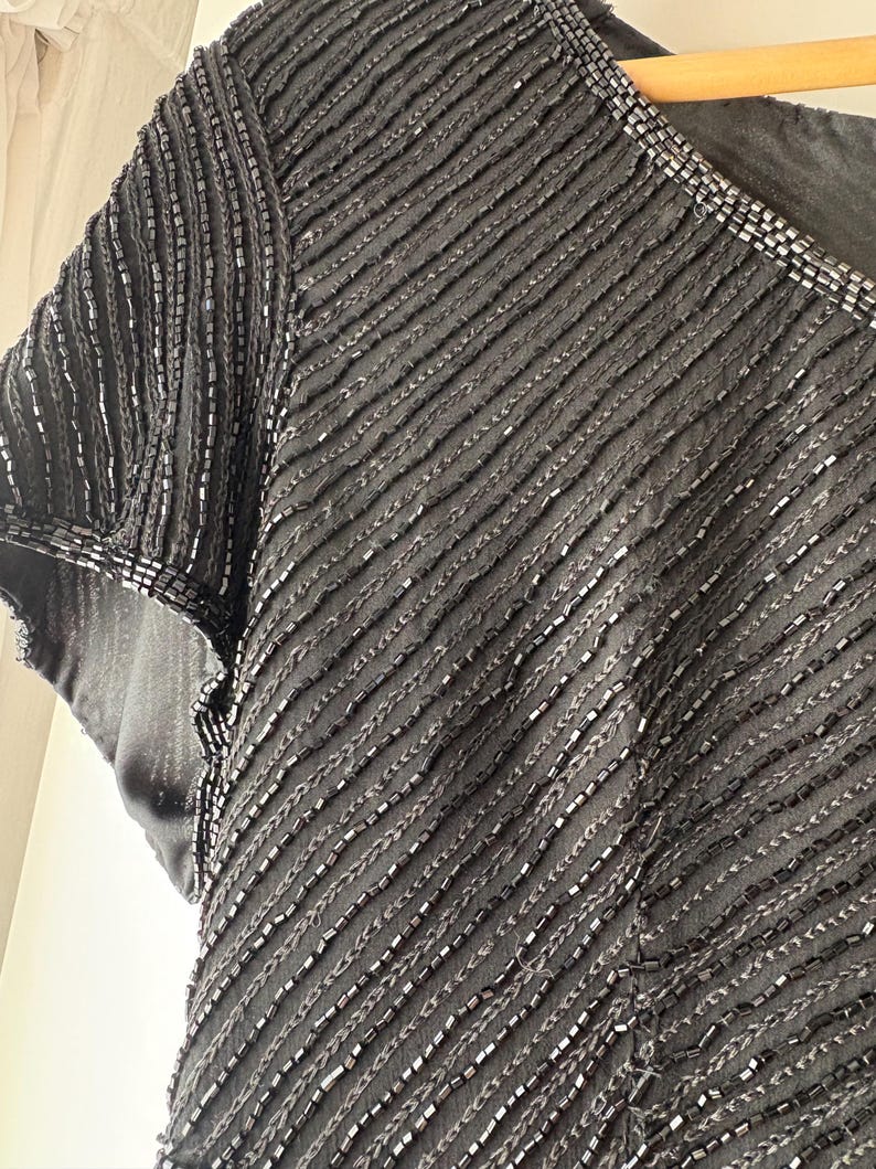 Peut inclure: Gros plan d'un haut noir &agrave; perles avec un d&eacute;collet&eacute; en V et des manches courtes. Le tissu est textur&eacute; avec des rang&eacute;es diagonales de perles, cr&eacute;ant un effet scintillant. L'encolure est &eacute;galement orn&eacute;e de perles.