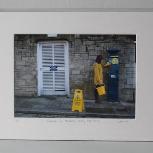 May include: A woman in a yellow raincoat uses a parking meter. A yellow caution sign is on the sidewalk. The text on the sign reads "Caution - Wet Floor".