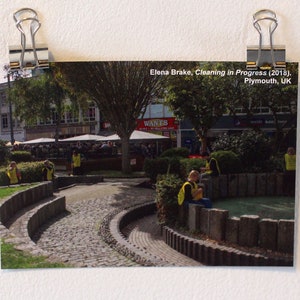 May include: A black and white photograph of a public square in Plymouth, UK. The photo is titled "Elena Brake, Cleaning in Progress (2018), Plymouth, UK". The photo shows a paved area with a circular stone fountain and a few people in yellow vests.