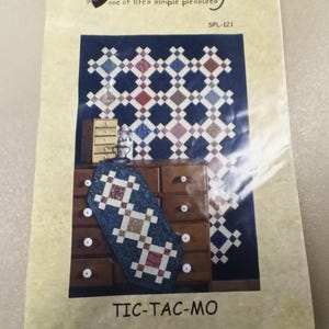 May include: A quilting pattern for a table runner or quilt called "Tic-Tac-Mo". The pattern features a blue background with red, white, and brown squares. The pattern is displayed on a wooden table with a drawer.