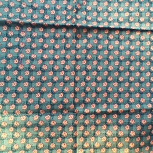 May include: Teal fabric with a repeating pattern of small, pink floral designs. The flowers are arranged in a grid pattern over a teal background with a subtle checkered texture. The fabric appears to be folded.
