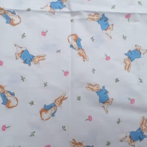 May include: White fabric featuring a repeating pattern of Peter Rabbit illustrations in various poses. The rabbits are depicted in their signature blue jackets and brown fur, interspersed with small pink flowers and green sprigs.
